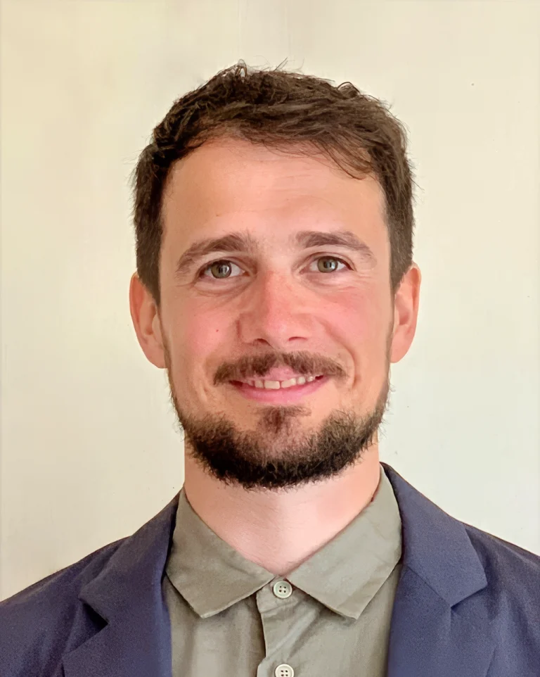 Smiling young man with short brown hair and beard wearing a blazer and button-up shirt, professional headshot.