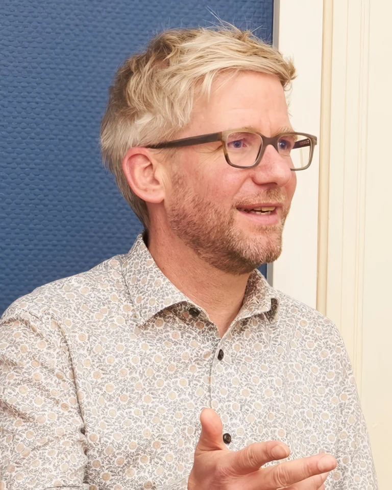 Blond man with glasses speaking and gesturing in a casual shirt.
